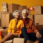 smiling senior couple sitting on floor with cardboard boxes and looking at camera, deciding whether to have an Estate Sales, Consignment, or Donation.